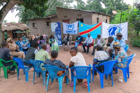 Outdoor community meeting with people sitting on blue chairs, facing a small stage with a banner.