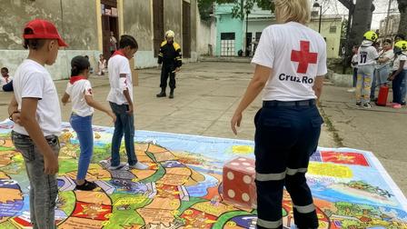 Red Cross volunteer with back turned beside a colorful game mat as children line up nearby.