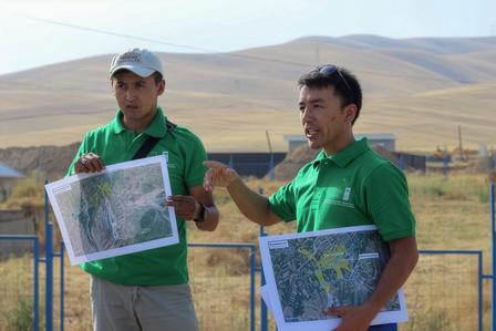 Two people in green shirts holding maps outdoors, one pointing in a rural landscape with hills.