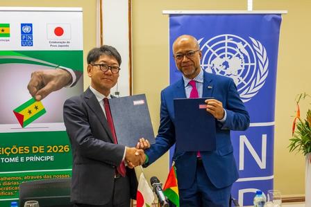 Two men in suits shake hands, each holding a document; UN banner and election poster in background.