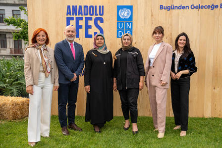 Seven people pose outdoors in front of a wooden wall with Anadolu signage and UN logos.