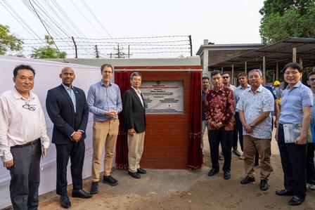 Group of men standing in a line in front of a small building with a white sign and red gate.