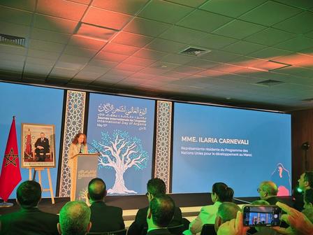 Conference stage with two blue screens and a central blue panel showing a tree; portrait and flag on the left; audience seated.