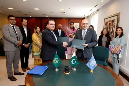 Two men exchange a document at a signing ceremony; officials stand nearby in a conference room.