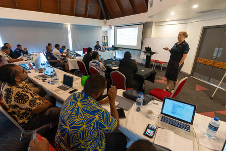 Photo of a workshop: attendees around tables while a presenter speaks at the front with a screen.