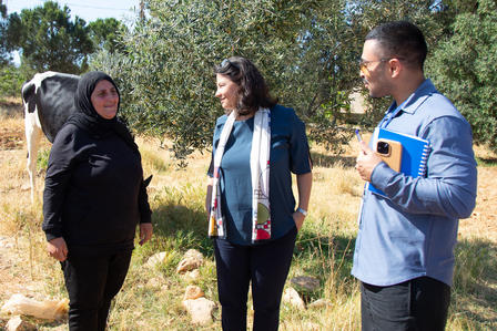 Three people stand in a sunlit field, talking; woman in hijab, others in casual clothes.