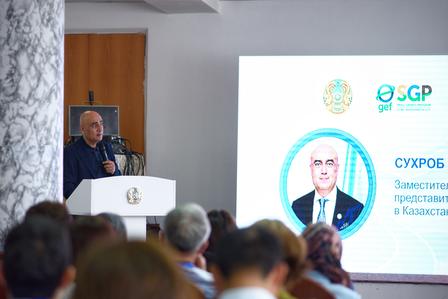 Speaker at podium addressing audience; slide shows circular logo with Cyrillic text.