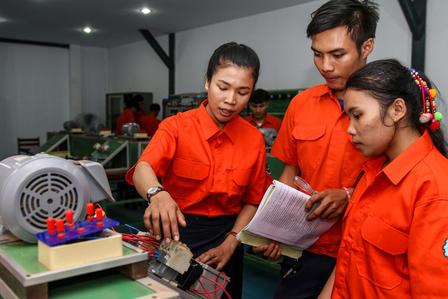 Three workers in orange uniforms examine an electronic board on a workbench in a workshop.