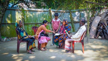 Group of women in colorful clothing seated in a circle on plastic chairs outdoors under trees.