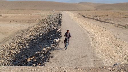 A lone cyclist pedals along a dusty desert road, with a rocky embankment to the left.