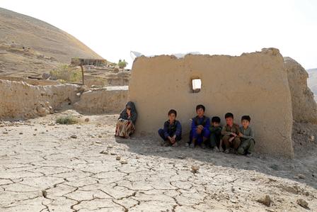 Group of people sitting by a crumbling mud-brick wall in a desert, cracked earth and distant hills.