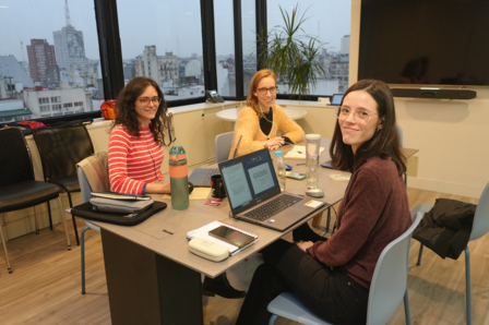 Photograph: three women at a table with laptops in a bright office overlooking the city.