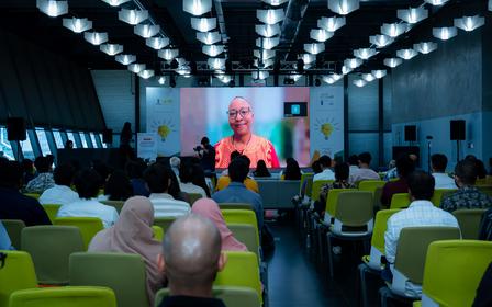 Photo: conference hall with colorful chairs; audience facing a large screen displaying a speaker.