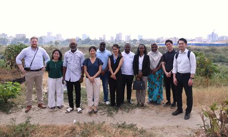 Group photo of diverse people posing outdoors on a dirt path with distant city skyline.