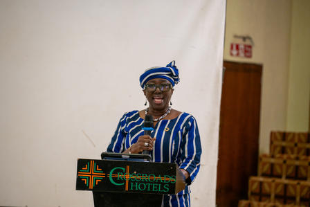 Person in blue striped outfit and hat speaks at podium; plain wall and chairs in background.