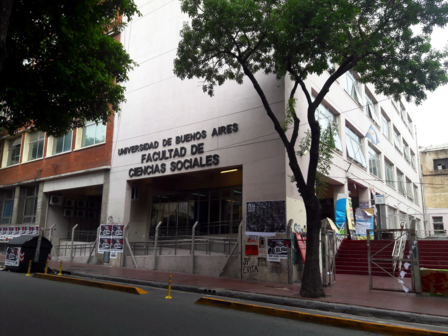 Tree-lined street in front of a white university building with a sign near the entrance.