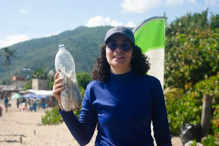 Person in blue shirt on sunny beach holding a plastic water bottle; green flag behind mountains.