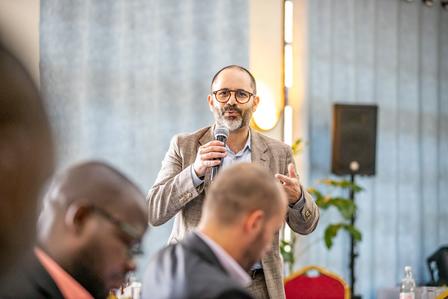 Person in a beige blazer takes a photo with a smartphone at a conference.