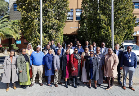 Large group of people posing outdoors, with trees and a building behind them.