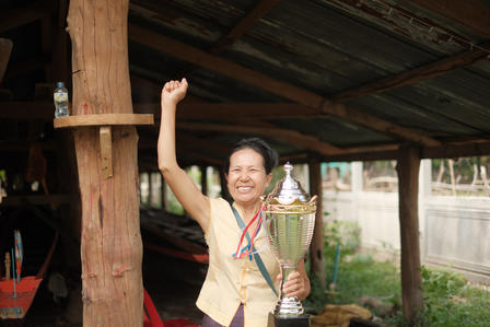 Smiling woman raises fist, holding a gold trophy and medal under a wooden shelter.