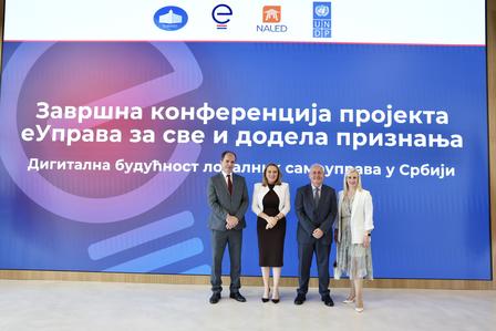 Photograph: Four people pose on a stage in front of a blue conference screen with logos.