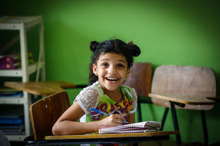 Photo of a young student at a desk in a classroom with green walls, writing.