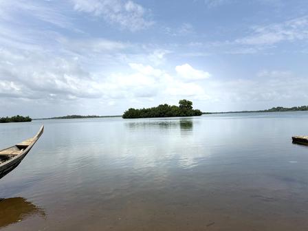 Tranquil lake with a small tree-covered island, partly cloudy blue sky, and boats along the shore.