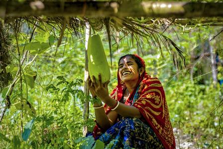 Woman in red-yellow patterned clothing sits among green foliage, hands in prayer; face blurred.