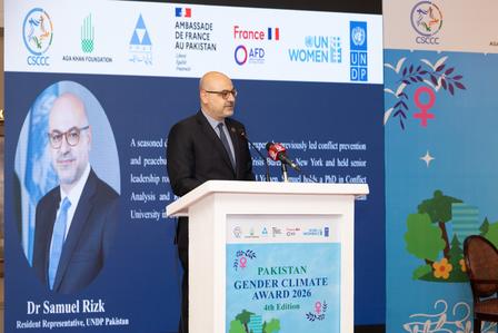 Photograph of a man in a suit speaking at a conference podium with blue banners.