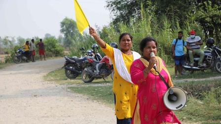 Two women with a megaphone and yellow flag on a dusty rural road; onlookers and bikes behind.