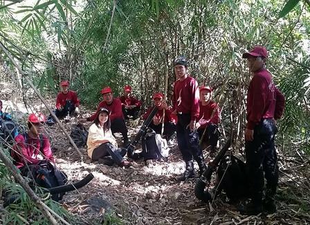 Group of people in red shirts gathered in a wooded clearing, some seated on the ground.