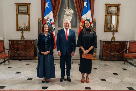 Group portrait of four people in formal attire standing in a grand room with flags in the background.