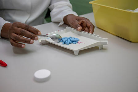 Hands weigh blue pills on a white tray with a scale, yellow container in the background.