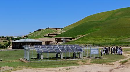 Hilly landscape with solar panels in a fenced area, a small building, and a group of people nearby.