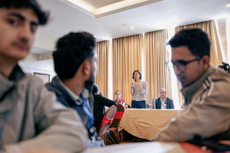 People in tan uniforms sit around a conference table as a speaker addresses a panel at the front.