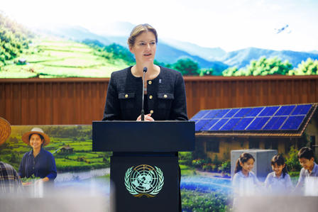 Photo: A woman speaks at a UN podium outdoors, mountains in the background.