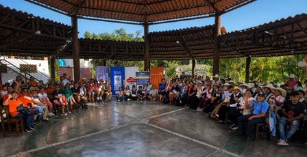Large crowd seated around a circular open-air wooden pavilion outdoors.