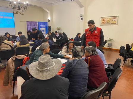 Community meeting; attendees sit around tables as a man in a red jacket speaks at the front.