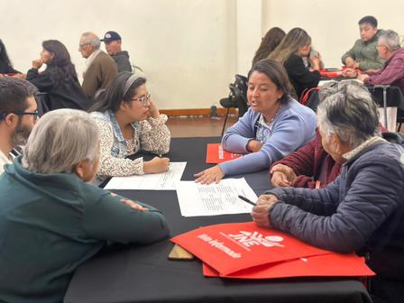 Diverse group of adults seated around a classroom table, discussing papers; red bags nearby.