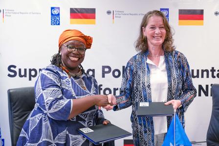 Two smiling women shake hands at a conference, each holding a folder, with banners in the background.