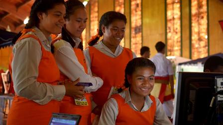 Photograph of girls in orange vests gathered around a computer in a warmly lit classroom.
