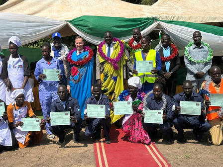 Group of people in colorful outfits holding certificates under a green tent and red carpet.
