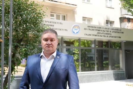 Man in a navy suit walks toward the camera outside a modern building with a glass entrance.