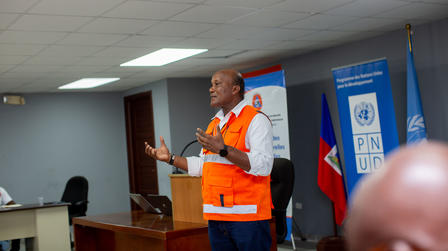 Photograph: Man in orange safety vest speaks at a podium in a conference room with UN banners.