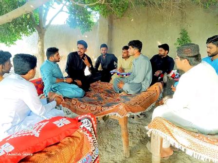 Group of men in traditional attire sitting in a circle around a low table outdoors under a tree.