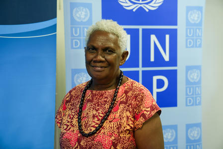 Photo: Smiling woman with short gray hair in a pink patterned blouse, in front of UNDP banners.