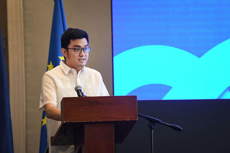 Man in white shirt speaks at a podium on stage; blue backdrop and flag nearby.