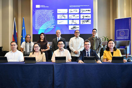 Group of people seated at a long blue table in a conference room, with a presentation screen.