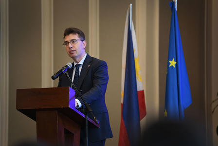 Photograph of a man in a suit speaking at a podium, with flags in the background.