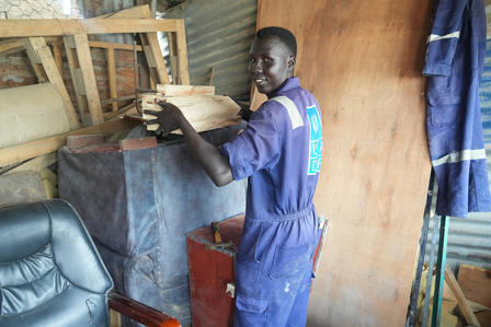 Photograph of a man in blue coveralls working on a wooden frame in a cluttered workshop.